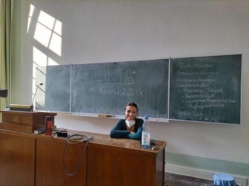 Lena bei einem Workshop in der Uni Bonn. Sie sitzt vorne am Pult, an der Tafel hinter ihr steht "FLINTA* in die Kommunalpolitik"