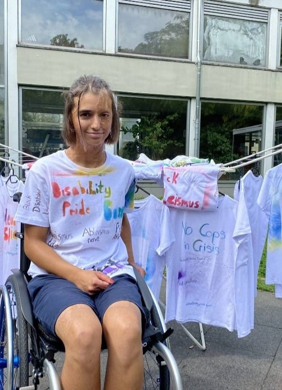 Lena im Rollstuhl sitzt vor einem Wäscheständer mit weißen T-Shirts, die besprayt sind. Lena hat auch ein "Disability Pride Bonn" T-Shirt an.