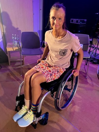 Lena bei einer Veranstaltung auf dem Podim, mit Rock, bunten Schuhen und im Rollstuhl